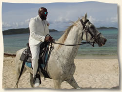 Groom in white suit on white horse riding to his beach wedding.
